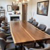 A modern dining room featuring a long wooden table with a natural grain, surrounded by eight gray chairs. The room has a contemporary chandelier, large framed artwork, a small cabinet, and a decorative vase near the white fireplace.