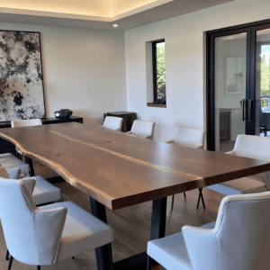 A modern conference room features a long wooden table with a natural edge, surrounded by eight white chairs. A large abstract painting and a lamp sit on a sideboard by the wall. Large glass doors reveal an outdoor view.