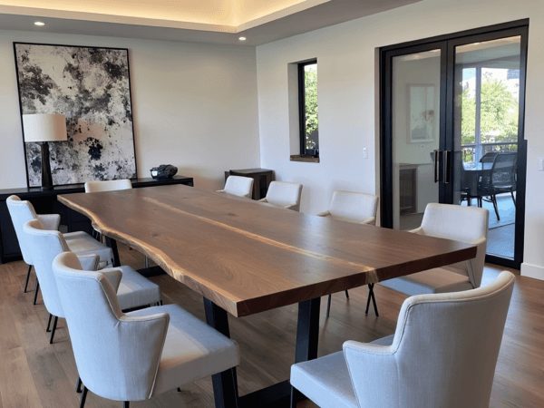 A modern conference room features a long wooden table with a natural edge, surrounded by eight white chairs. A large abstract painting and a lamp sit on a sideboard by the wall. Large glass doors reveal an outdoor view.