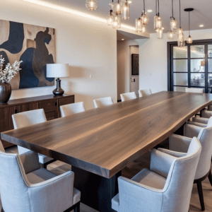 A modern dining room with a long wooden table surrounded by cushioned chairs. A sideboard with a lamp and vase with white flowers is against the wall. Abstract art hangs above, and several pendant lights are suspended from the ceiling.