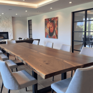 A modern dining room with a large wooden table surrounded by white upholstered chairs. A fireplace is visible in the background, and a colorful painting hangs on the wall. Large windows provide natural light and a view of greenery outside.