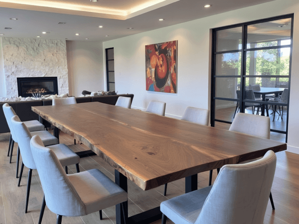 A modern dining room with a large wooden table surrounded by white upholstered chairs. A fireplace is visible in the background, and a colorful painting hangs on the wall. Large windows provide natural light and a view of greenery outside.