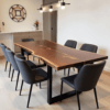 A modern dining room features a live edge wooden table with black metal legs, surrounded by six dark gray upholstered chairs. A minimalist light fixture hangs above, and a wall with a decorative mirror and vase adds a touch of elegance.